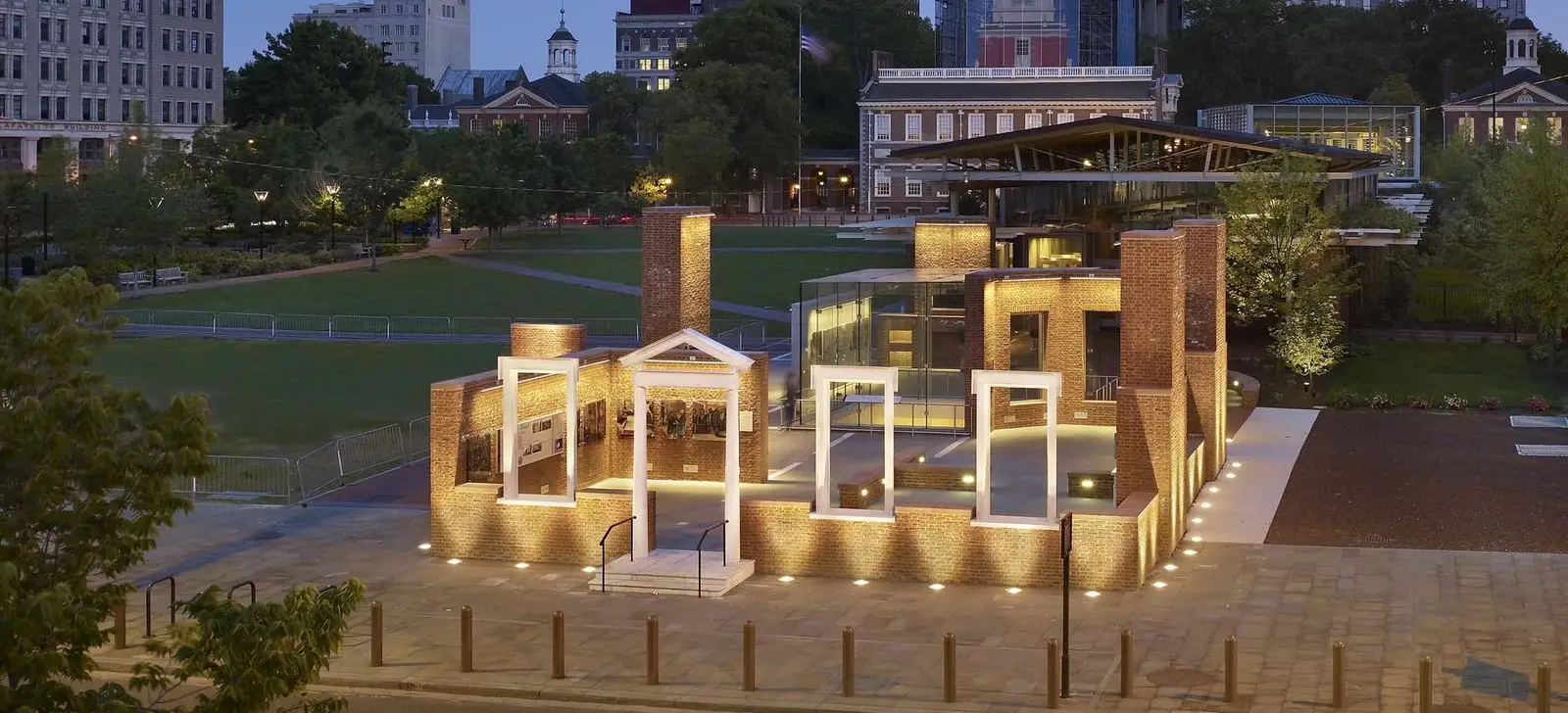 An illuminated outline of a brick civic building at dusk, with open door and window frames, set in a park area surrounded by trees and historic buildings in the background.