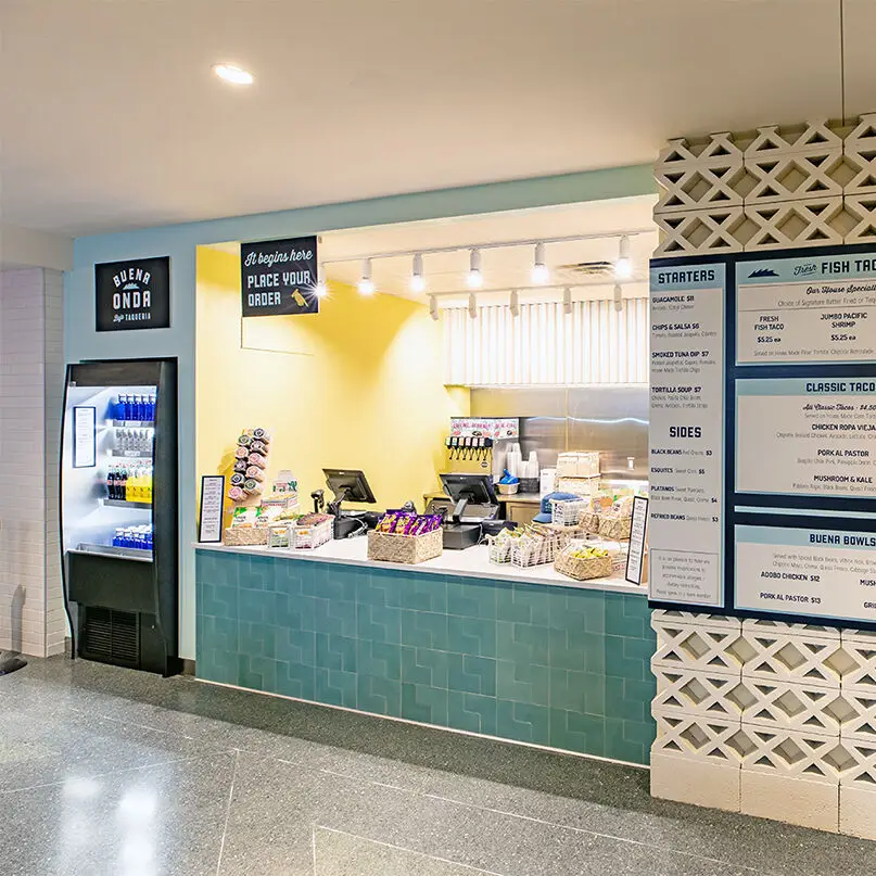 A modern food counter with a teal tiled front showcases thoughtful interior design, snacks and drinks on display, a wall menu, a Place Your Order sign overhead, and a refrigerated drink cooler to the left.