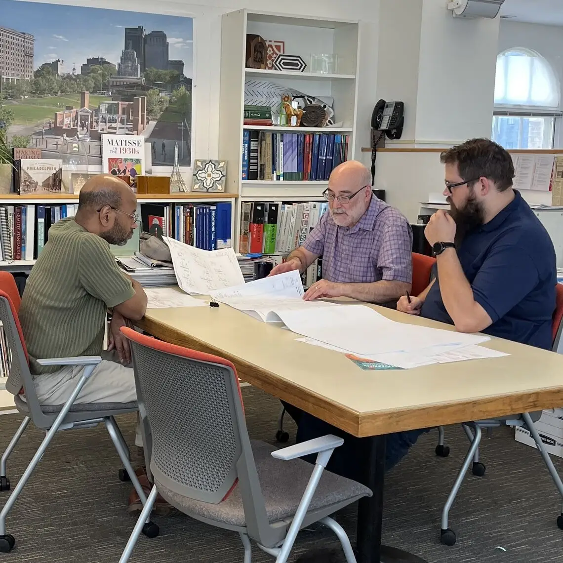 Three men sit around a table in an office, reviewing large architectural blueprints related to historic preservation. Bookshelves and a cityscape photo are in the background. The mood is focused and collaborative.