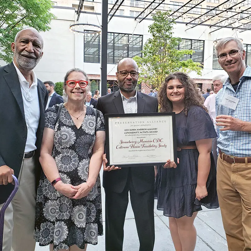 Five people stand together at an outdoor event in Philadelphia. The man in the center, an architect, holds a large framed award certificate. All are smiling and dressed in business or casual attire, with other guests and a pergola visible in the background.