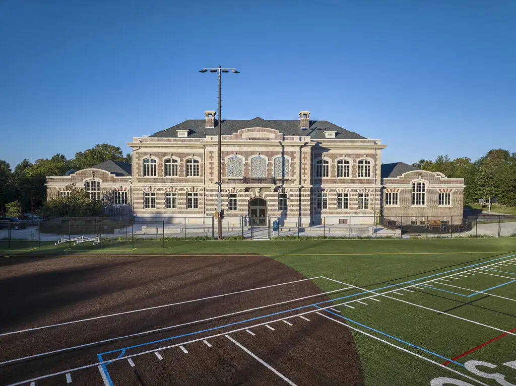 A historic, multi-story stone building with arched windows and intricate detailing, set behind a civic sports field with green turf and white painted lines under a clear blue Philadelphia sky.
