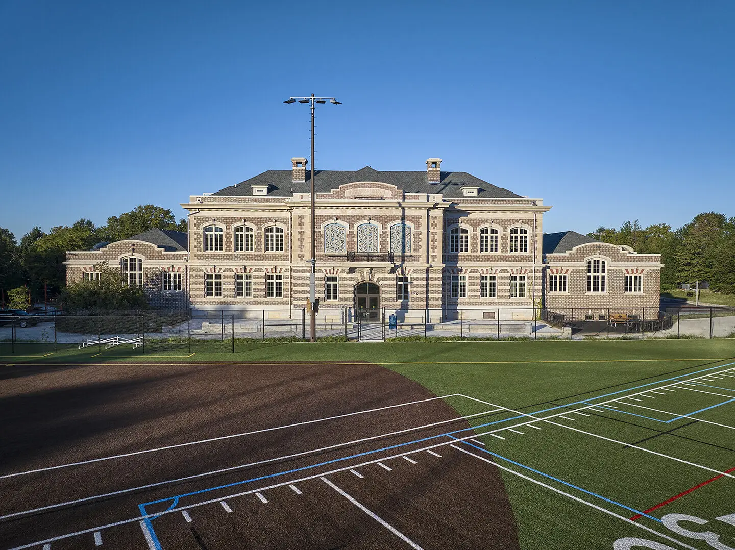 A historic, multi-story stone building with arched windows and intricate detailing, set behind a civic sports field with green turf and white painted lines under a clear blue Philadelphia sky.