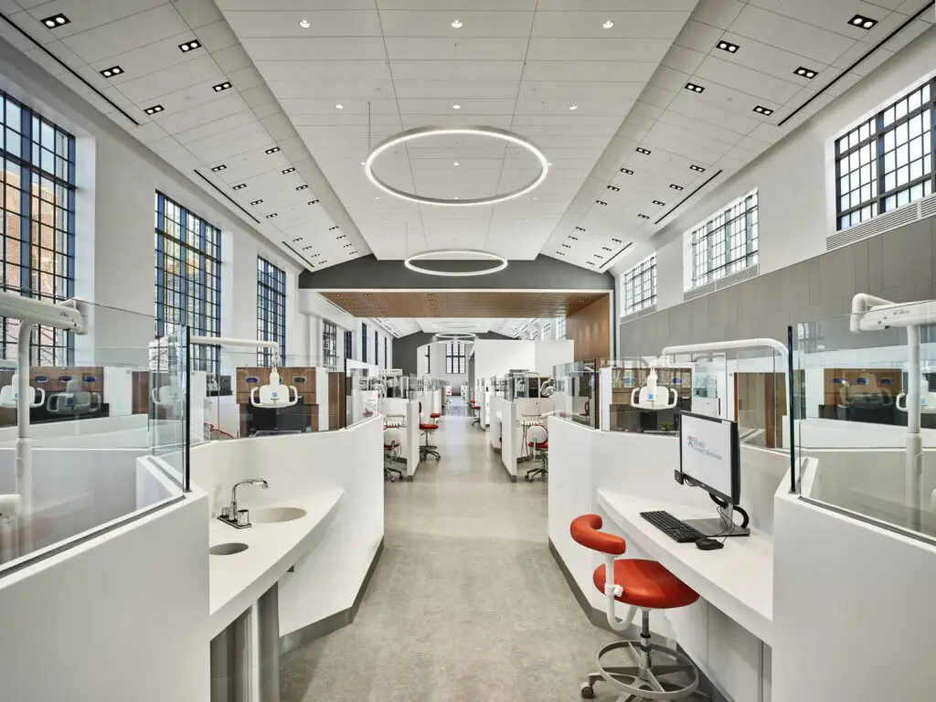 A modern, bright dental clinic featuring expert interior design with white workstations, red chairs, computers, and glass partitions. Large windows line the walls, while circular ceiling lights illuminate the spacious, clean interior.
