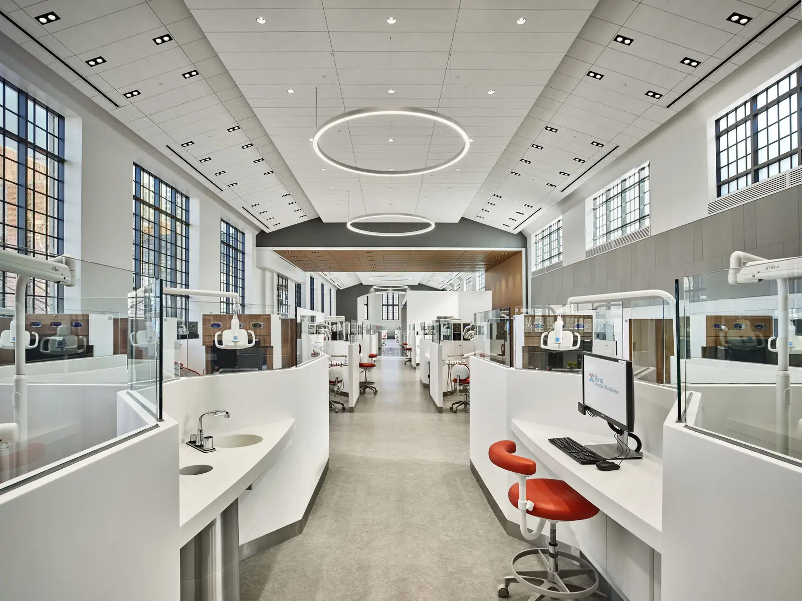 A modern, bright dental clinic featuring expert interior design with white workstations, red chairs, computers, and glass partitions. Large windows line the walls, while circular ceiling lights illuminate the spacious, clean interior.