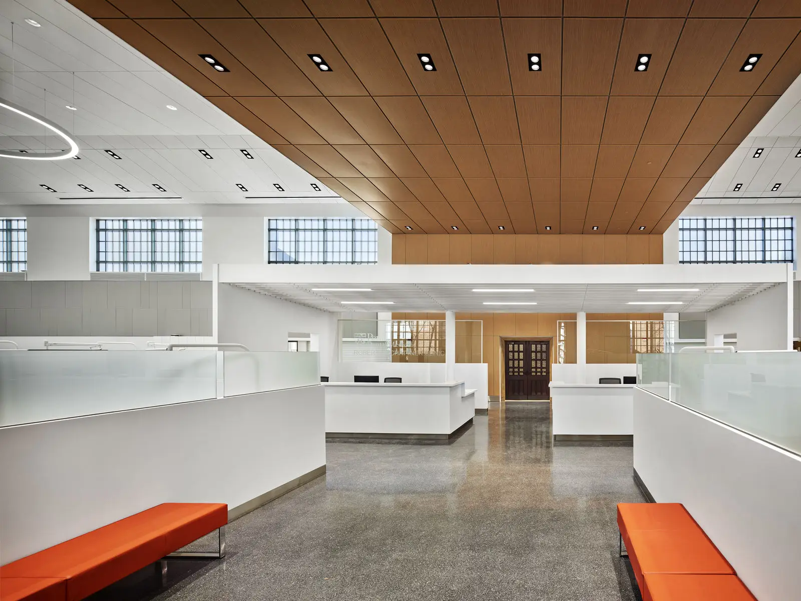 A modern, spacious office lobby with white walls, frosted glass partitions, polished concrete floors, orange benches, and a wooden ceiling panel evokes a civic atmosphere. Large windows let in natural light. Reception desks are visible in the background.