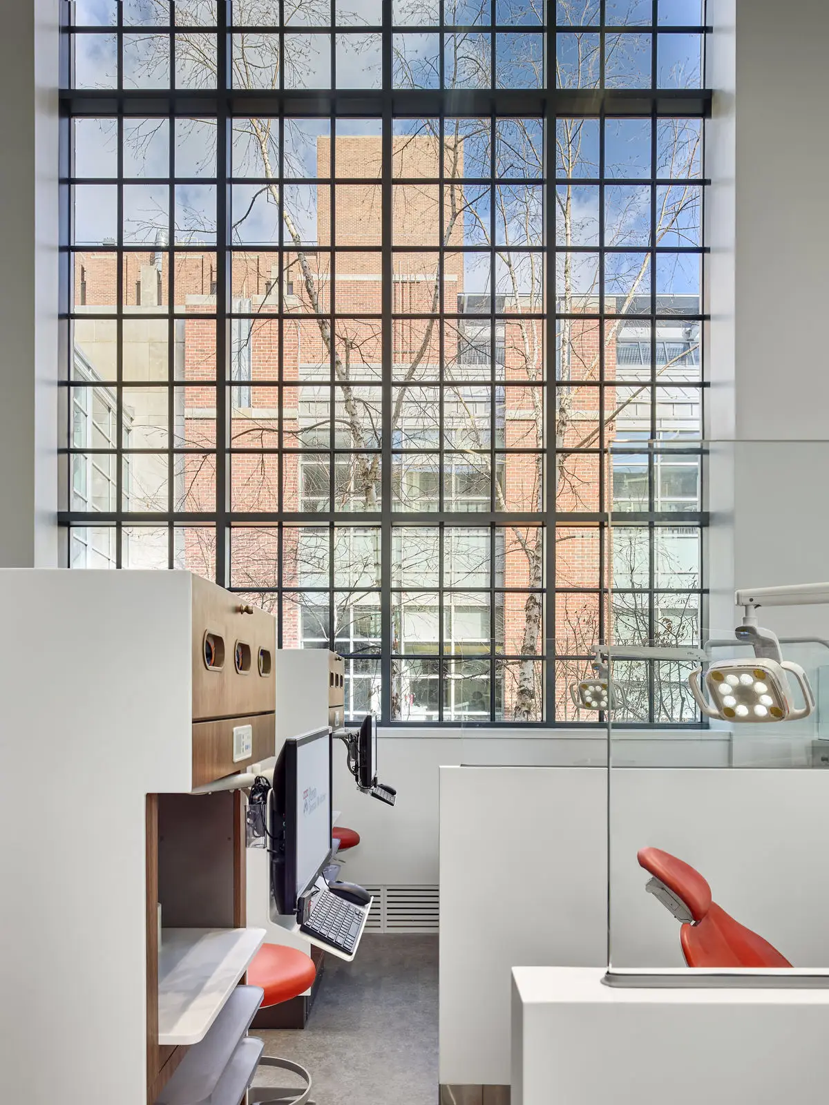 A modern dental clinic in Philadelphia with red dental chairs and computer workstations, set against a large window with a grid pattern, showcasing stylish interior design and views of trees and a brick building outside in daylight.