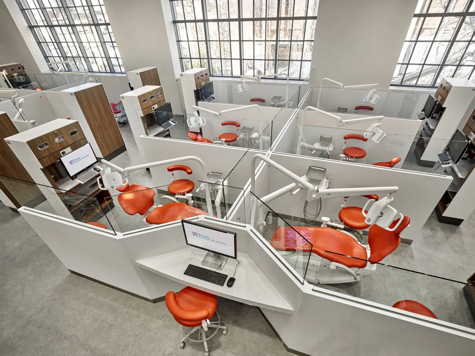 A modern dental clinic in Philadelphia with educational resources, multiple workstations featuring orange chairs, dental equipment, glass dividers, and computers. Large windows fill the well-designed interior with natural light, highlighting the clean, organized space.