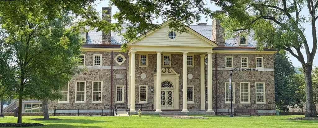 A large, historic stone building with tall columns, symmetrical windows, and a triangular pediment serves as a civic landmark, surrounded by green grass and trees under a clear sky.