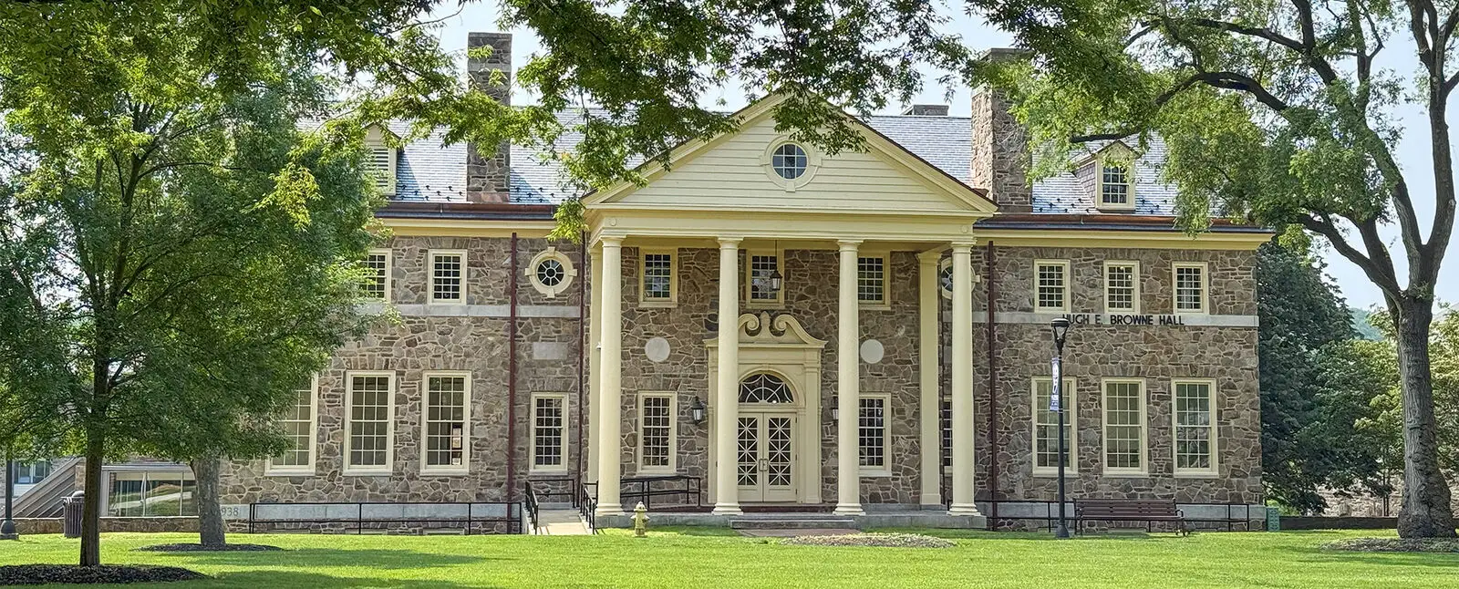 A large, historic stone building with tall columns, symmetrical windows, and a triangular pediment serves as a civic landmark, surrounded by green grass and trees under a clear sky.