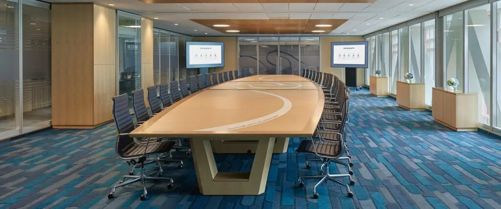 A modern conference room designed by an architect features a long wooden table, black swivel chairs, large windows, blue-patterned carpet, two wall-mounted screens, and decorative plants near the windows.