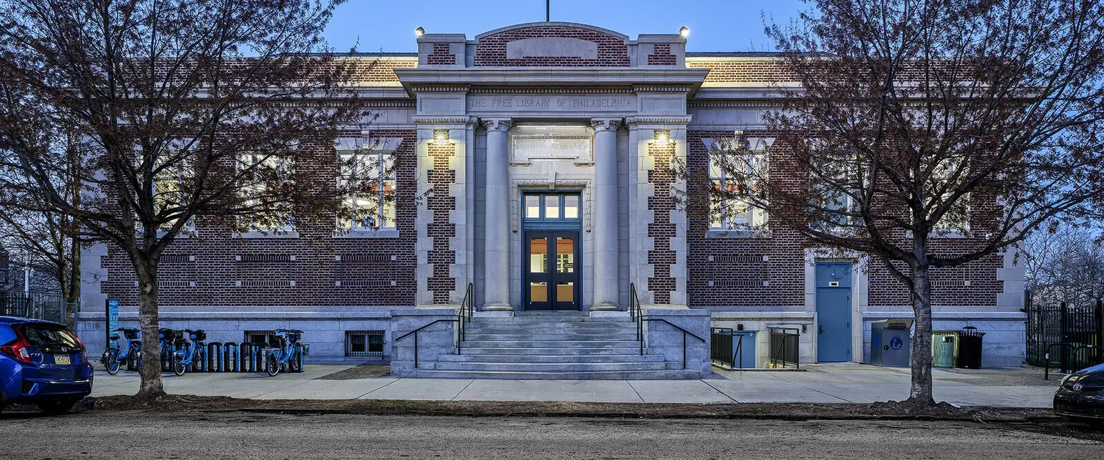 A civic brick building with tall columns, stone steps, and decorative architectural details—showcasing historic preservation—glows at dusk. Leafless trees frame the entrance, and bicycles are parked to the left.