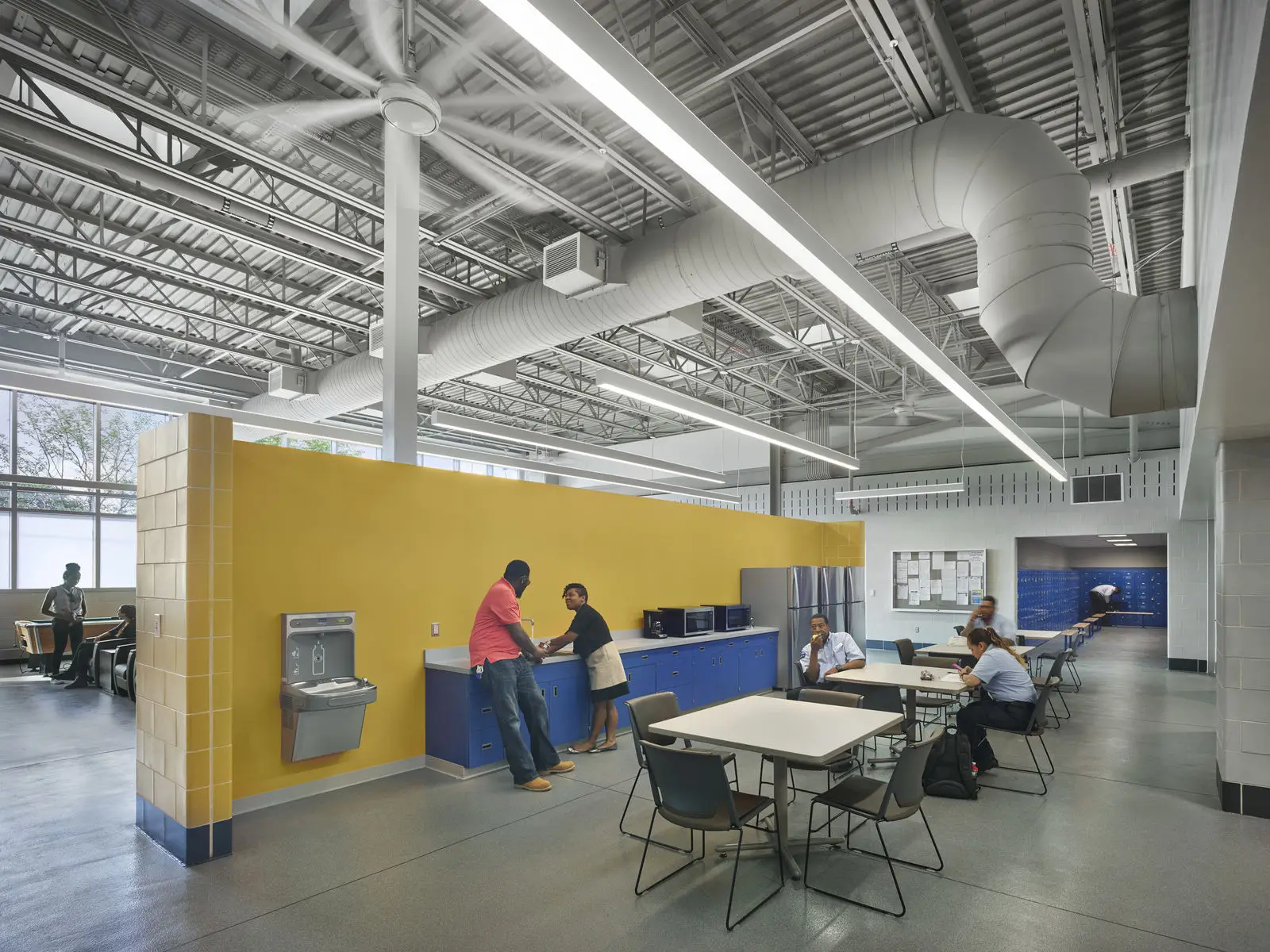 A modern break room designed by an architect features yellow and white walls, exposed ceiling ducts, tables, and chairs. Several people are sitting, talking, or using the kitchenette near blue cabinets. Large windows provide natural light.