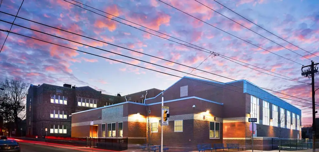 A modern brick school building in Philadelphia is seen at sunset, its windows glowing with light. The colorful sky and overhead power lines create a civic scene, while a street sign and leafless tree stand in the educational setting’s foreground.