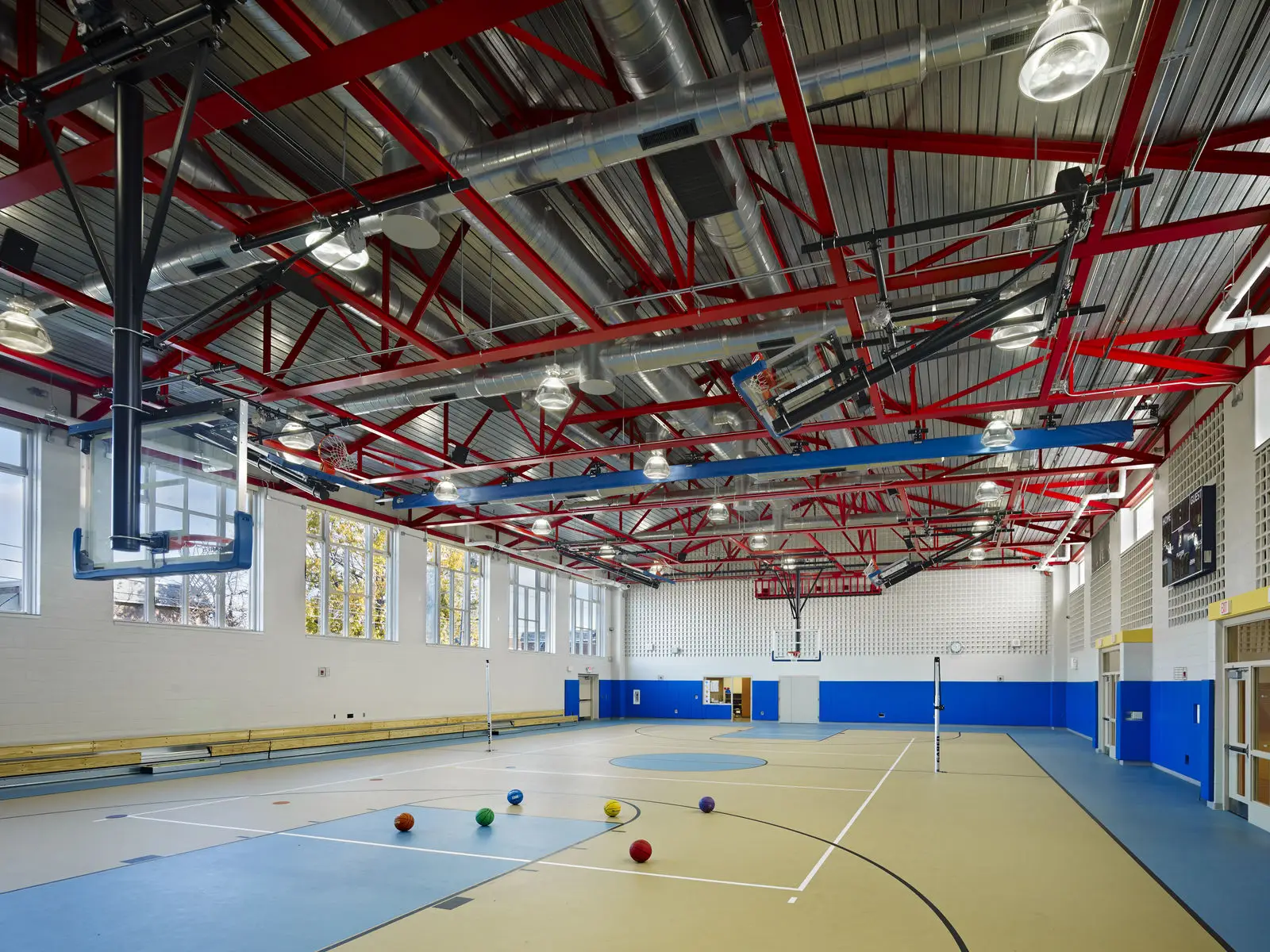 A spacious indoor gymnasium with a high ceiling, red exposed beams, and thoughtful interior design by a talented architect. Basketball hoops and colorful balls scatter the tan and blue court while large windows brighten bleachers along one wall.