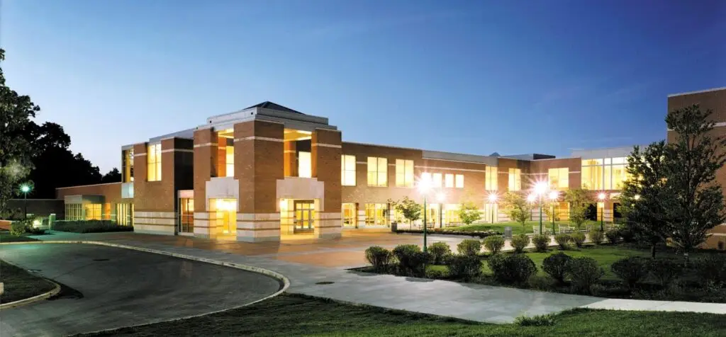 A modern, two-story brick building with large windows is brightly lit at dusk. The curved driveway in front is bordered by landscaped shrubs and trees, blending educational purpose with striking interior design; the sky is clear and blue.