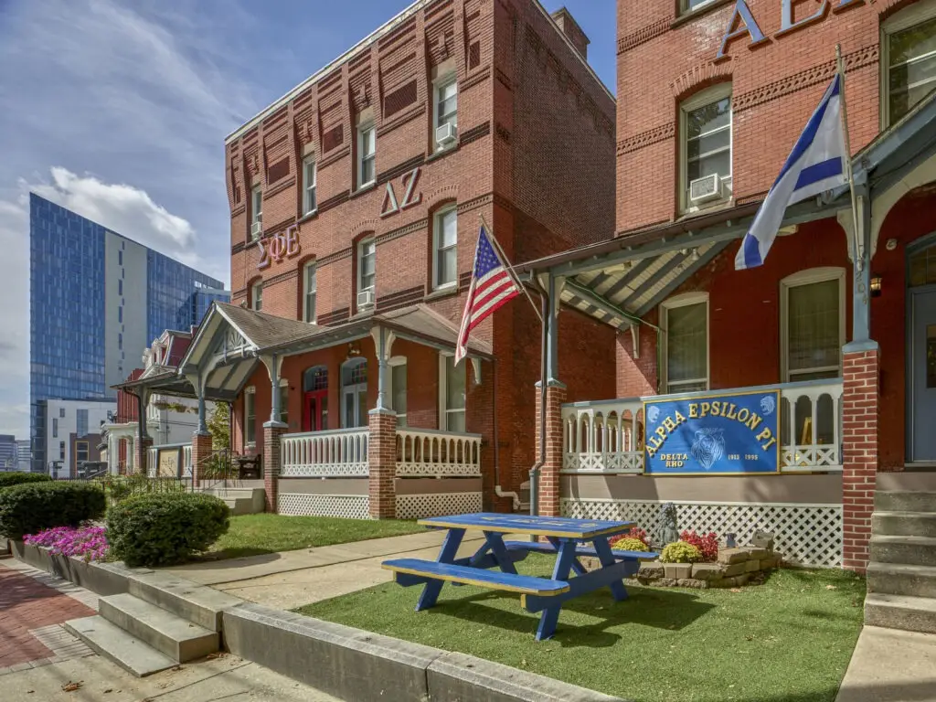 A red brick fraternity house with Greek letters, educational banners, American and Greek flags, a blue picnic table on the lawn, and modern buildings in the background highlights both tradition and campus life.