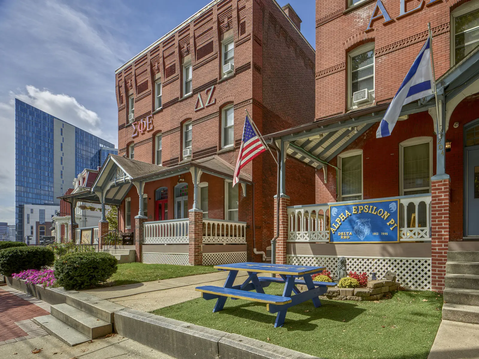 A red brick fraternity house with Greek letters, educational banners, American and Greek flags, a blue picnic table on the lawn, and modern buildings in the background highlights both tradition and campus life.