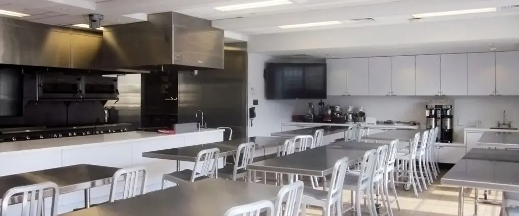 A bright, modern commercial kitchen classroom in Philadelphia with stainless steel tables and chairs, white cabinets, kitchen appliances, and large cooking ranges—an inviting space for educational culinary experiences.