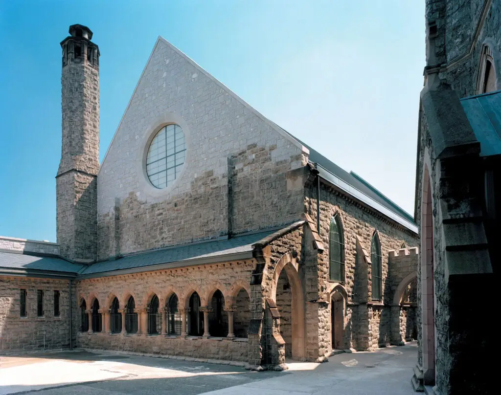 A historic stone building with arched windows, a circular window on a steep gable, and a tall cylindrical chimney against a clear blue sky, showcasing gothic-style arches and textured stone walls—an excellent example of historic preservation.
