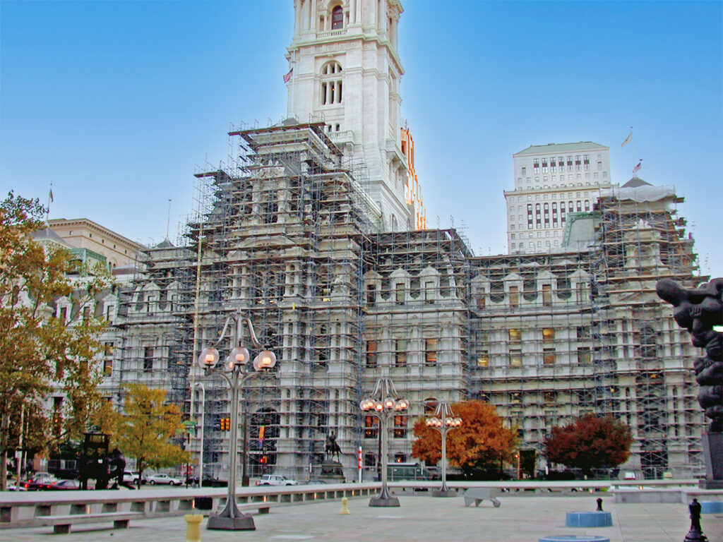 Philadelphia City Hall covered in scaffolding for restoration, surrounded by autumn trees and lit street lamps, with tall buildings in the background against a clear blue sky.
