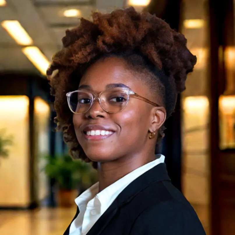 A person with short, natural curly hair, wearing glasses, a white shirt, and a black blazer, smiles in a brightly lit, modern indoor setting—an architect surrounded by blurred plants and lights that suggest an educational environment.