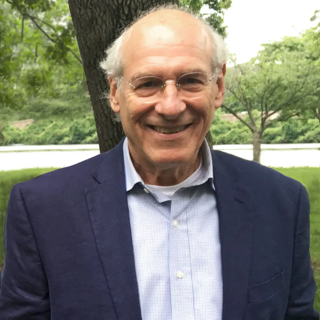 An older man with glasses and a friendly smile stands outdoors in Philadelphia, wearing a blue blazer and light blue shirt. Green foliage, a body of water, and his passion for civic engagement and historic preservation shine in the background.