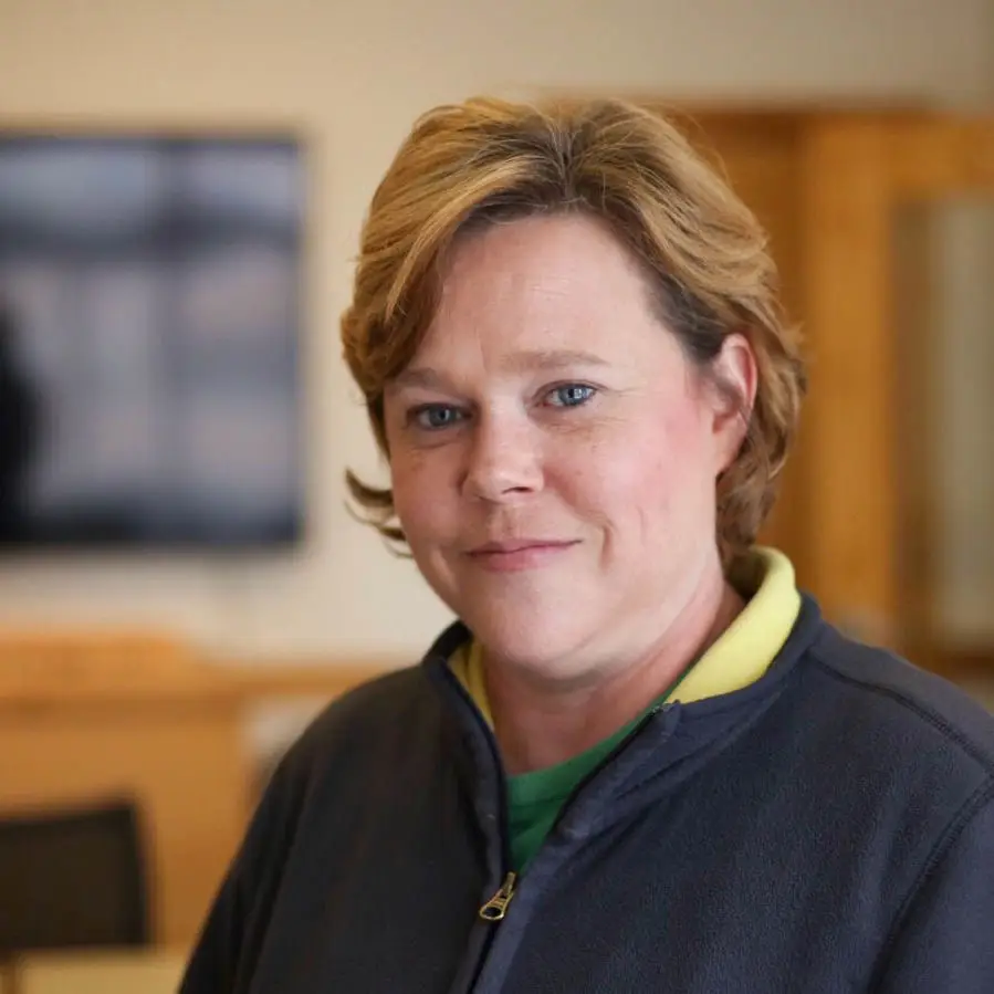 A woman with short light brown hair and a blue zip-up jacket stands indoors, softly smiling at the camera. The blurred background, featuring a TV screen and wooden shelves, hints at her interest in interior design.