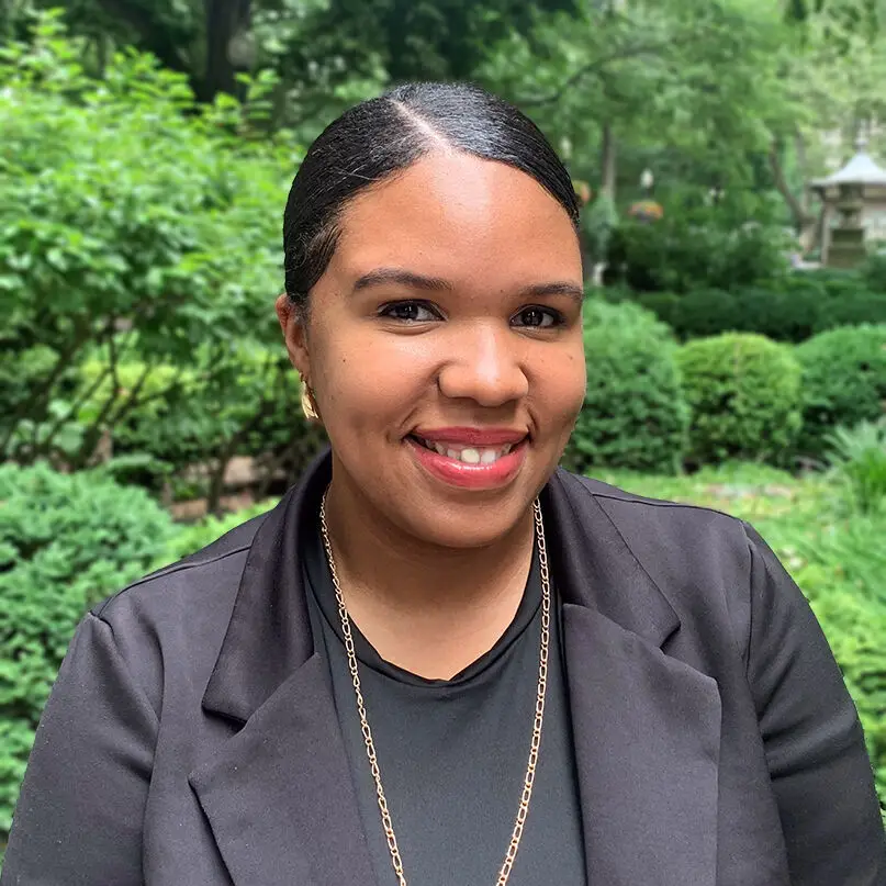 A woman with dark hair pulled back, wearing a black blazer, black shirt, and gold necklace, smiles outdoors in a green, leafy garden or park setting in Philadelphia.