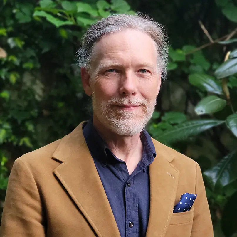 A middle-aged man with gray hair and a beard, dressed in a brown jacket, navy shirt, and polka-dot pocket square, stands outdoors by green leafy plants. The Philadelphia architect smiles slightly at the camera.
