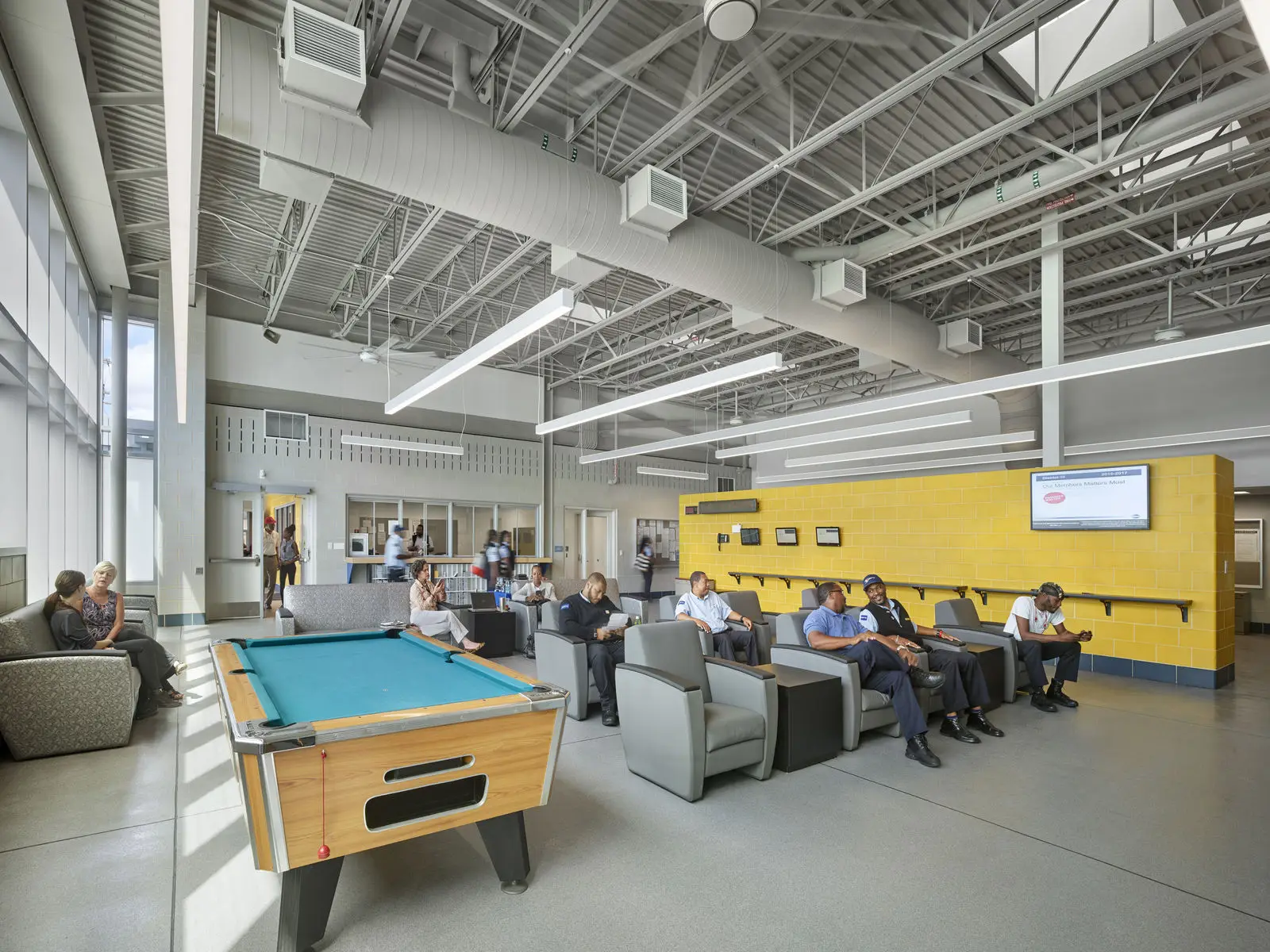 A spacious, modern lounge in Philadelphia with a pool table, several people sitting in armchairs, and a bright yellow accent wall. Large windows let in natural light, and there is a TV mounted on the wall—perfect for educational gatherings.