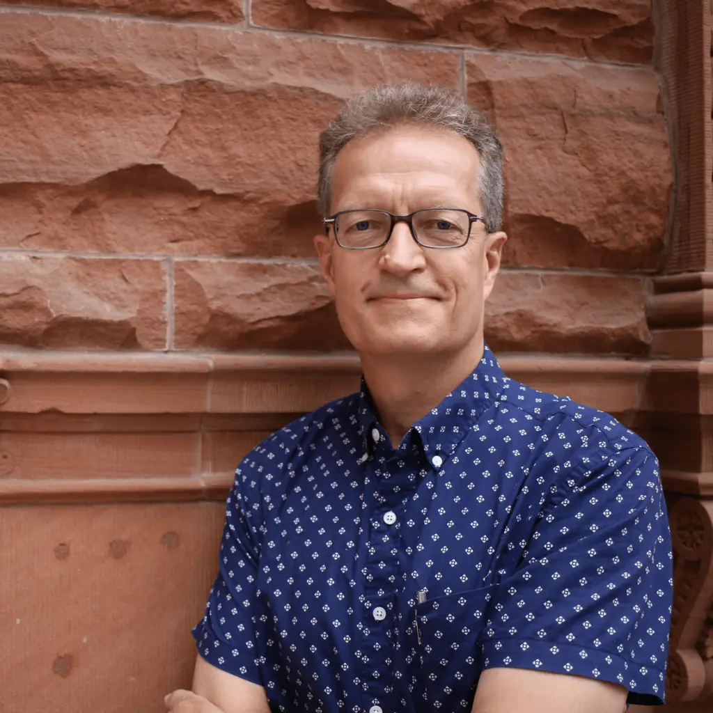A Philadelphia architect with short gray hair and glasses stands in front of a red stone wall, wearing a navy blue patterned short-sleeve shirt and smiling slightly with his arms crossed, reflecting his passion for historic preservation.