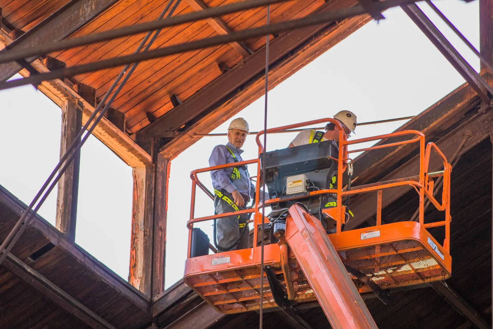 Two construction workers in safety gear stand on an orange lift, working inside a wooden structure with exposed beams and an open roof. Bright daylight illuminates this Philadelphia civic project in progress.