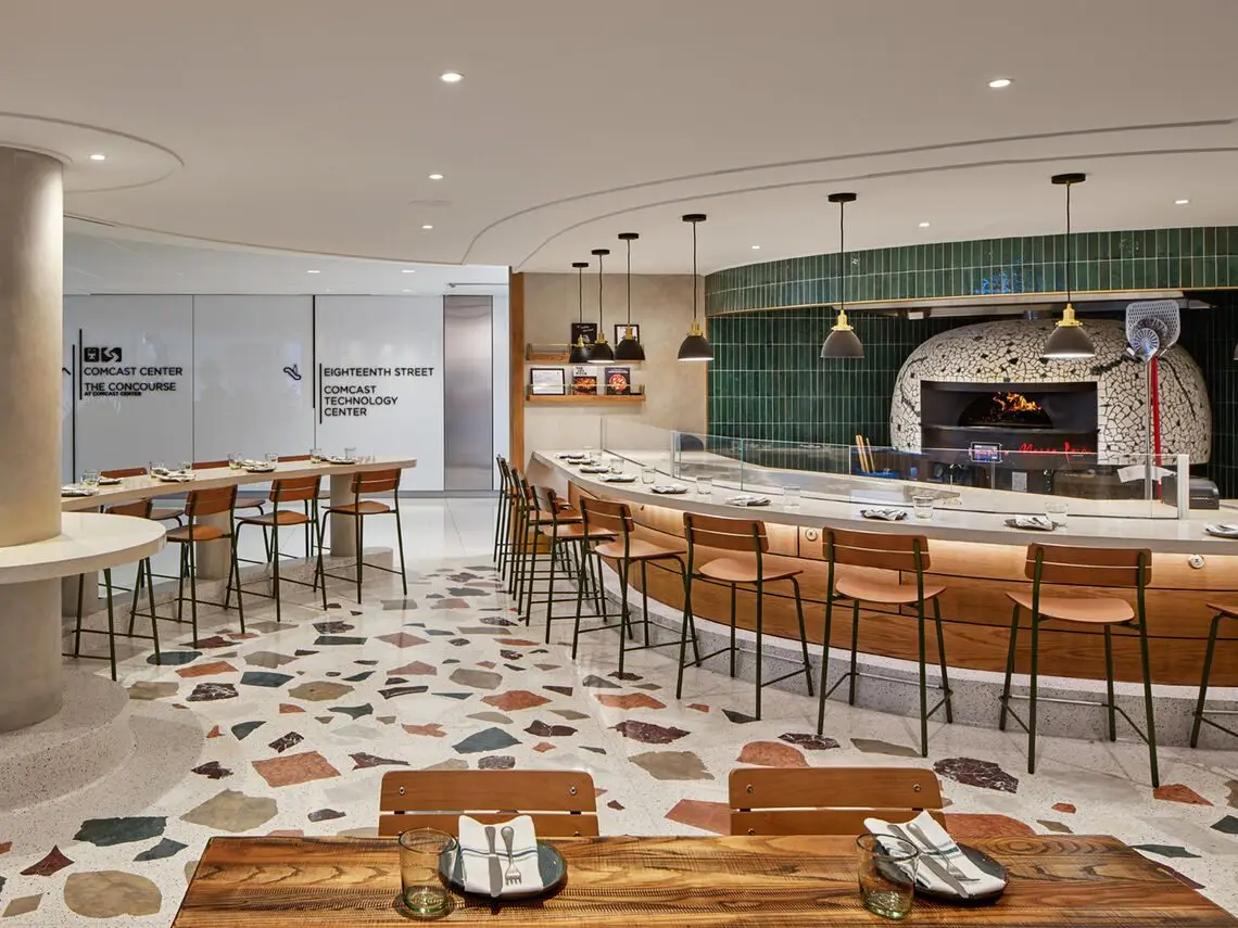Modern restaurant interior design in Philadelphia with a curved bar featuring wooden stools, an open pizza oven with green tile backdrop, terrazzo flooring, and a communal wooden dining table. Glass doors and signage are visible in the background.