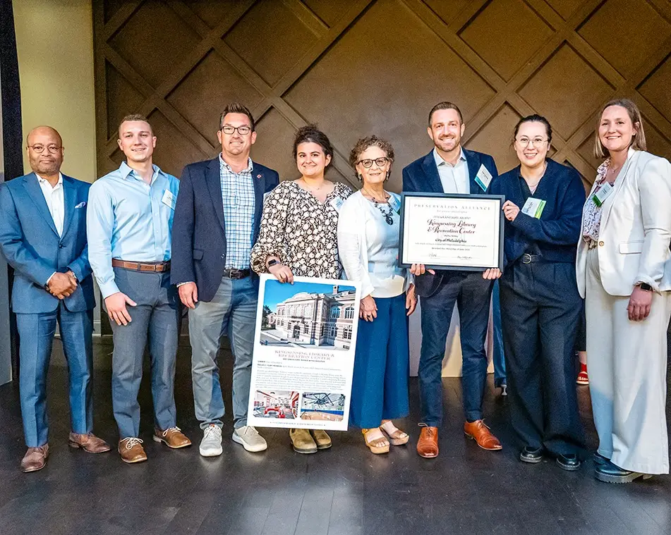 Eight people stand in a row, smiling at the camera; two in the center hold awards, including a large framed certificate and a poster featuring Kingsessing Library and Recreation Center as KMA wins Preservation Alliance award, with a geometric wall background behind them.