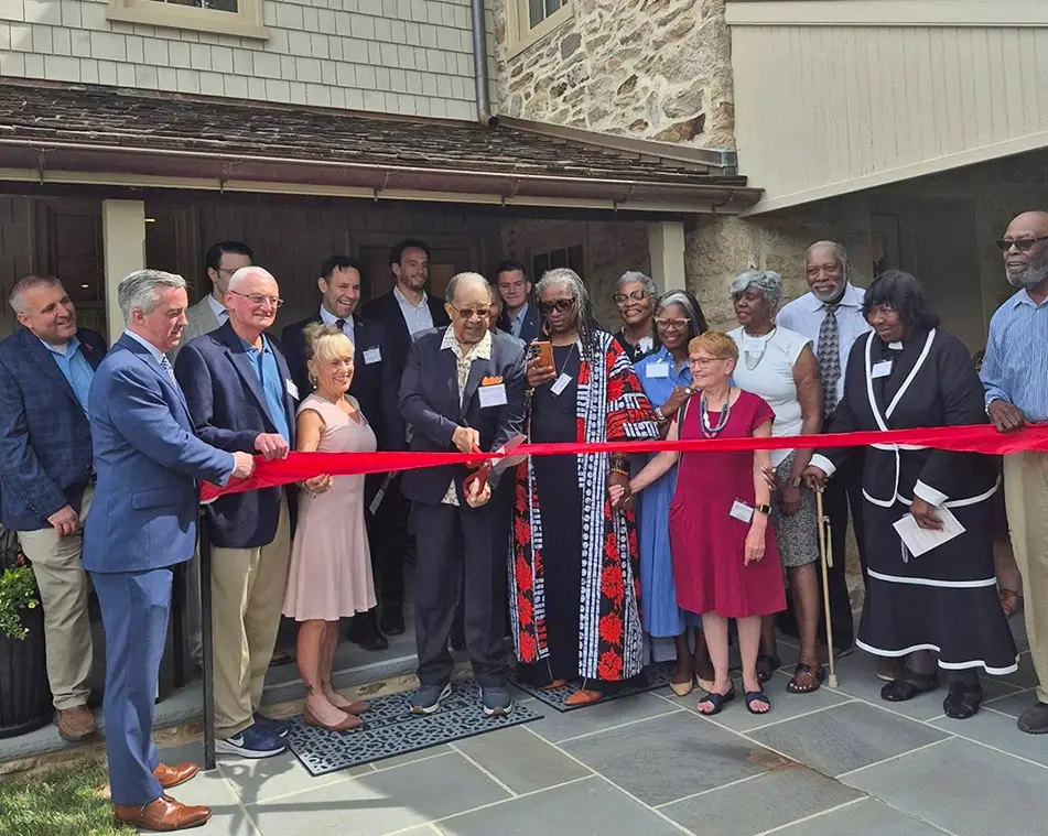 A diverse group of people stand together outside a stone building, smiling as they cut a large red ribbon during a ribbon-cutting ceremony. Some hold the ribbon, others pose for the photo.