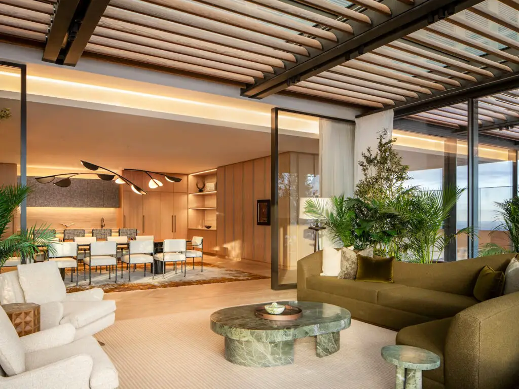 Modern living and dining area with large glass doors, green plants, light-colored armchairs, a green sofa, and a marble coffee table, featuring wood accents and soft lighting.