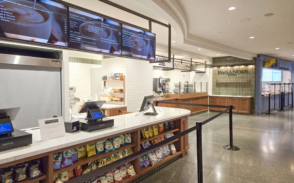 Modern cafe counter with digital menus, cash registers, snacks on display, and a tiled white and wood interior. No customers or staff are visible. Black stanchions guide the queue.