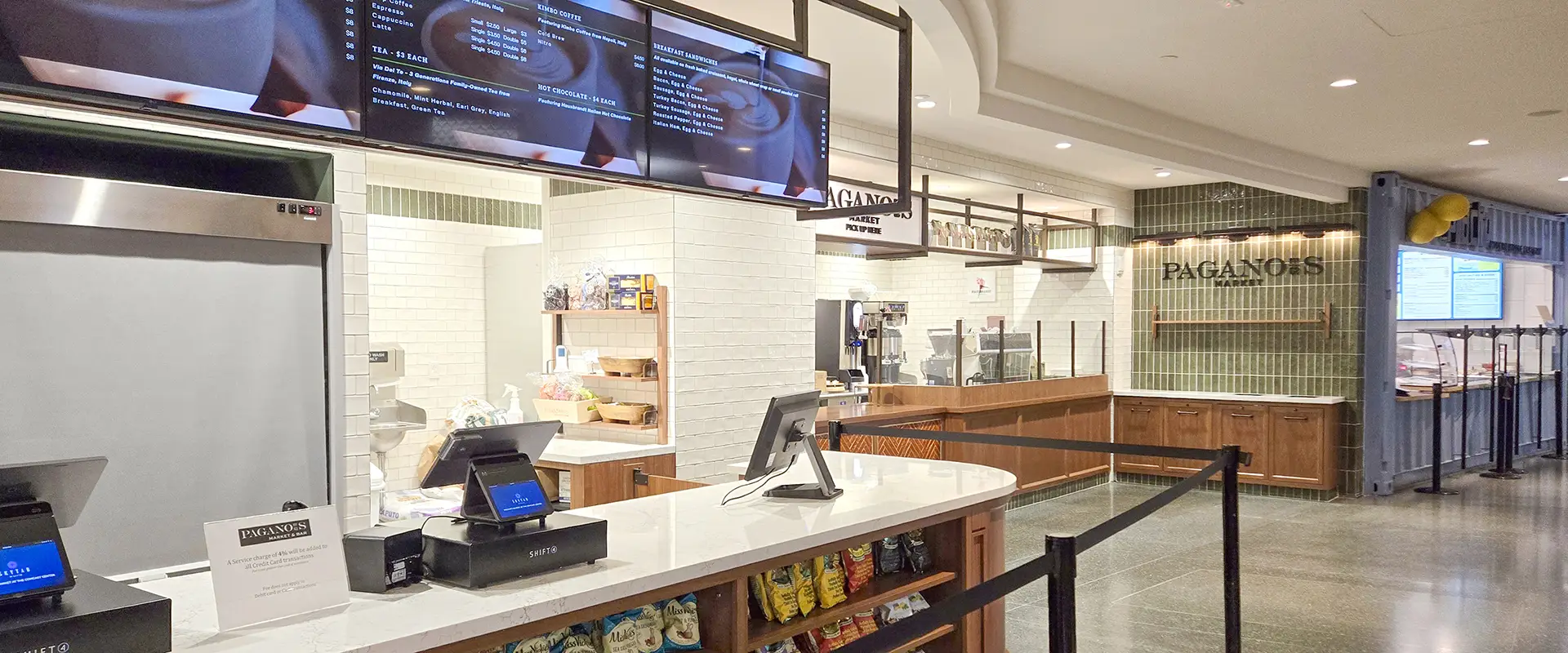 A clean, modern counter-service restaurant with digital menu screens, cash registers, and a white-tiled kitchen behind the counter. The name Paganos is visible on the wall in the background.