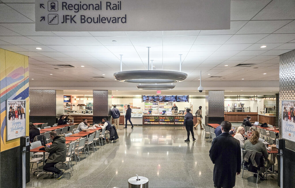Cafeteria with people sitting at tables and others walking; menu boards and food counter in the background. Overhead sign points to Regional Rail and JFK Boulevard.