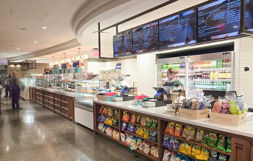 A modern cafeteria with a counter displaying snacks, drinks, and a menu screen overhead. A person stands at the register, and shelves are lined with chips and packaged foods. Another person is seen walking in the background.