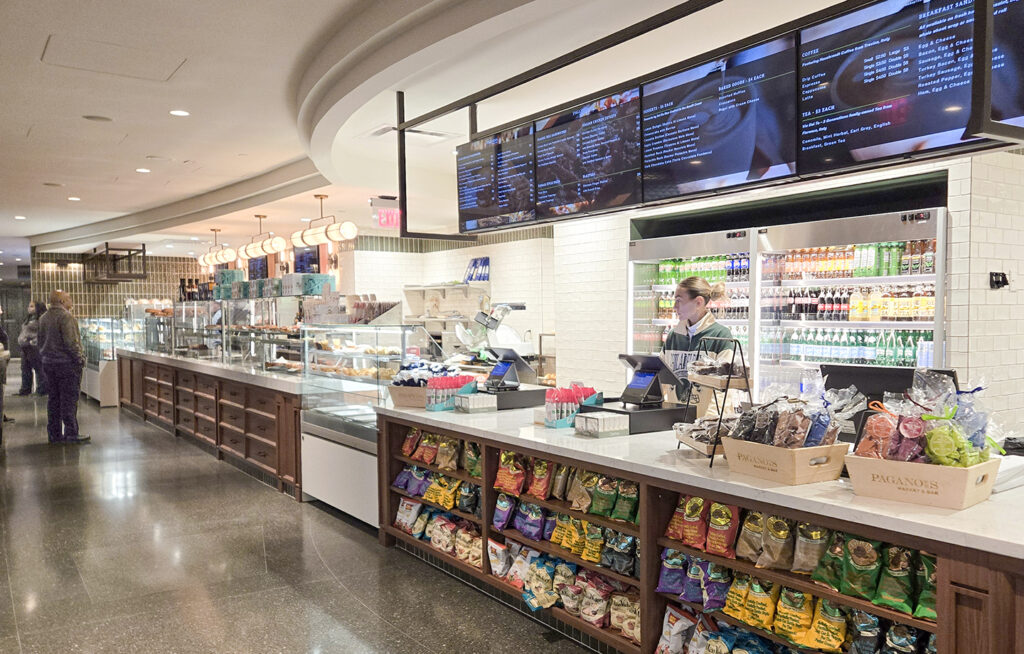 A modern cafeteria with a counter displaying snacks, drinks, and a menu screen overhead. A person stands at the register, and shelves are lined with chips and packaged foods. Another person is seen walking in the background.