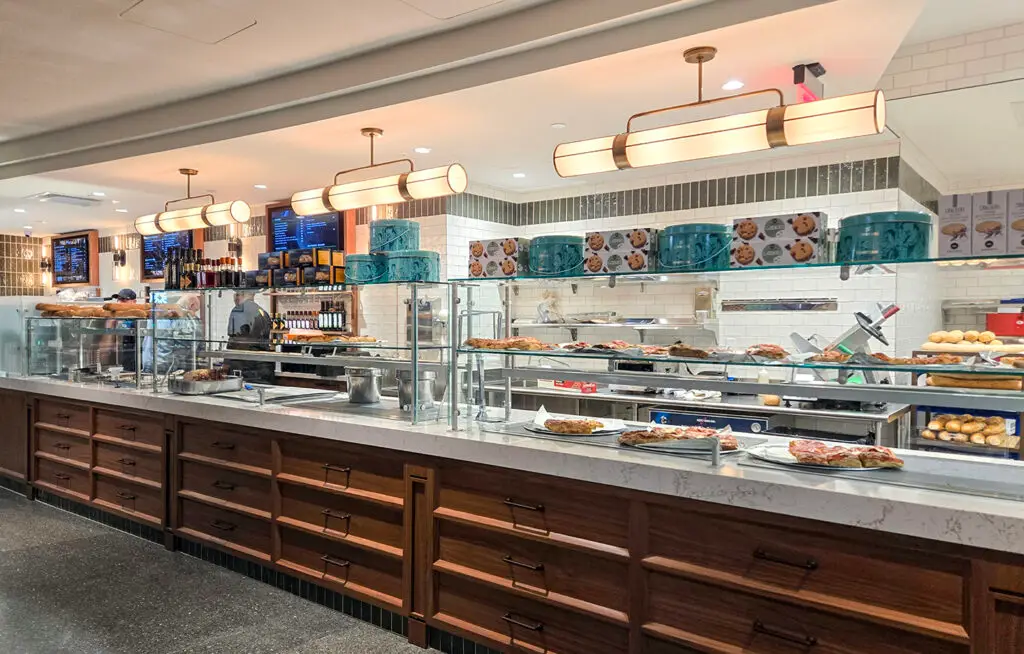 A well-lit cafeteria counter displays various foods under glass, with trays of baked goods and entrees. Blue trays stack on top, and pendant lights hang above the white countertop. Menus are visible in the background.