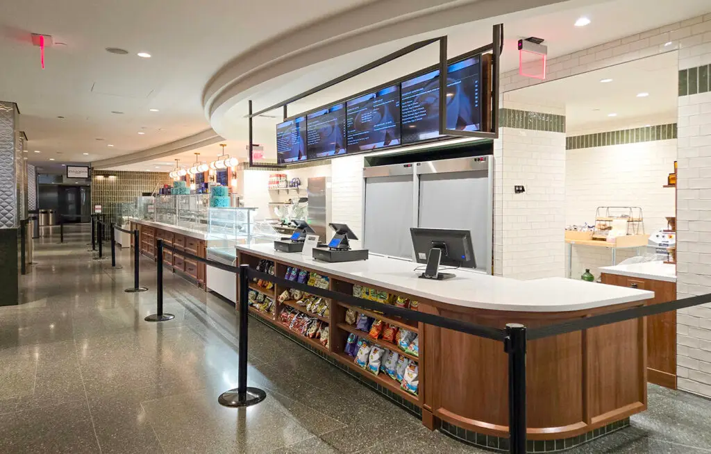 Modern cafeteria counter with digital menu boards, snack shelves, registers, and a glass food display. The area is clean, with black queue barriers guiding customers.