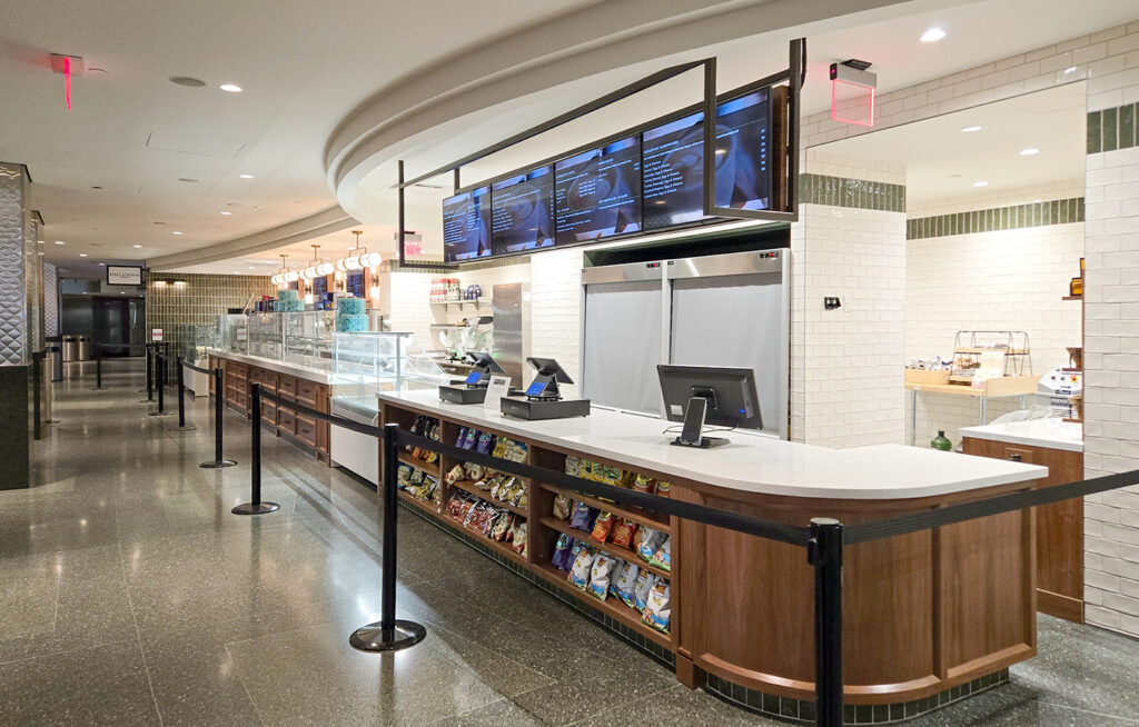 Modern cafeteria counter with digital menu boards, snack shelves, registers, and a glass food display. The area is clean, with black queue barriers guiding customers.