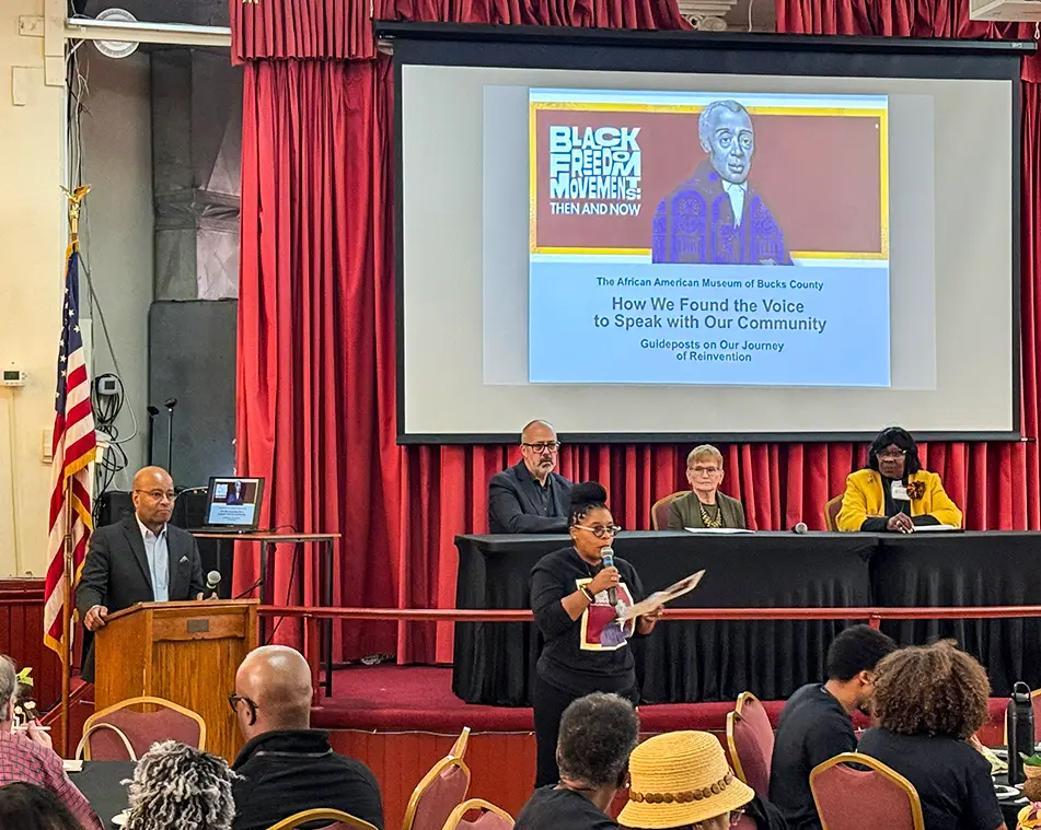 A panel discussion is underway in a large room with red curtains. A screen displays event info for the African American Museum of Bucks County. People sit at tables, and a woman stands speaking into a microphone.